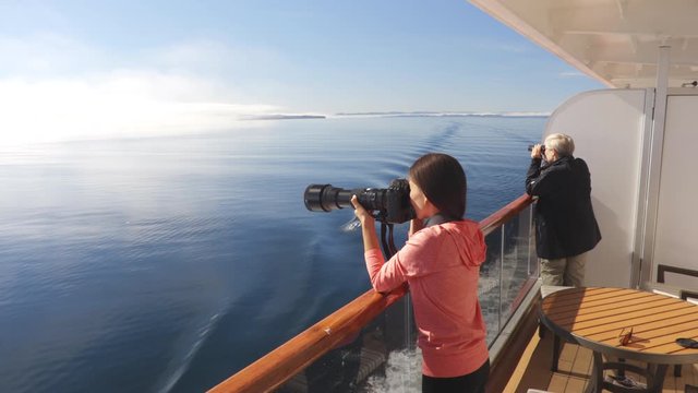 Alaska Glacier Bay Tourists looking at landscape from ise ship private balcony suite stateroom cabin using binoculars and taking pictures. People on vacation travel looking at wildlife cruising.