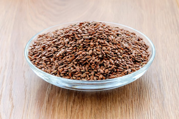 Heap of whole flax seeds in a glass saucer on a wooden table. Healthy food for lower cholesterol and preventing heart diseases.