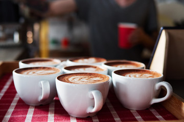 Set of freshly brewed cappuccino cups with milk foam latte art served on white red checkered tray in cozy city cafe lit with natural window light