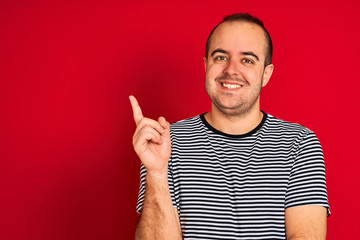 Naklejka premium Young man wearing striped navy t-shirt standing over isolated red background with a big smile on face, pointing with hand and finger to the side looking at the camera.