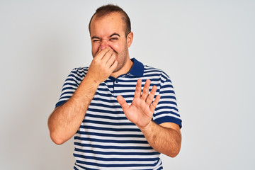 Young man wearing casual striped polo standing over isolated white background smelling something stinky and disgusting, intolerable smell, holding breath with fingers on nose. Bad smells concept.