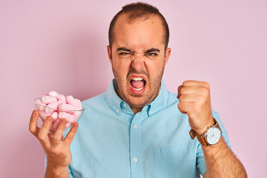 Young man holding bowl with marshmallows standing over isolated pink background annoyed and frustrated shouting with anger, crazy and yelling with raised hand, anger concept