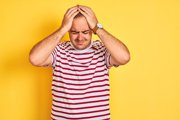 Young man wearing casual striped t-shirt standing over isolated yellow background suffering from headache desperate and stressed because pain and migraine. Hands on head.