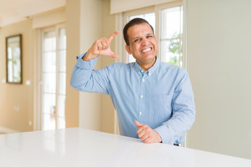 Middle age man sitting at home smiling and confident gesturing with hand doing size sign with fingers while looking and the camera. Measure concept.