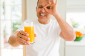 Middle age man drinking a glass of orange juice at home with happy face smiling doing ok sign with hand on eye looking through fingers