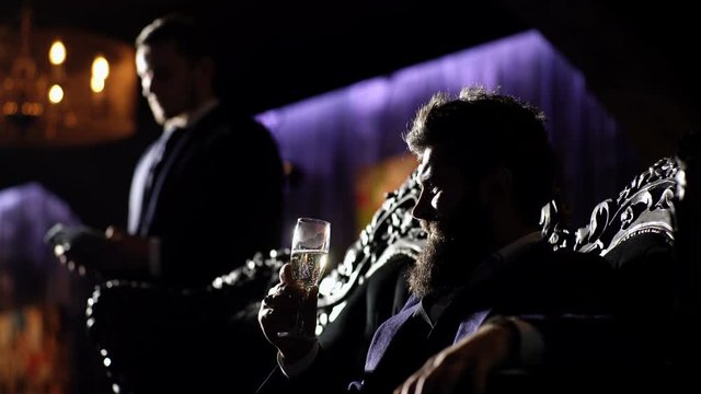 Bearded man with serious face and security man. Criminal boss in luxury suit sits on vintage sofa. Mature man with beard talks to his assistant. Crime, mafia, gangster concept. Luxury rich lifestyle.