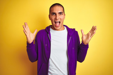 Young handsome man wearing purple sweatshirt over yellow isolated background celebrating crazy and amazed for success with arms raised and open eyes screaming excited. Winner concept