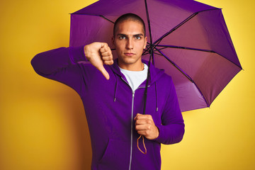 Young handsome man wearing purple umbrella over yellow isolated background with angry face,...