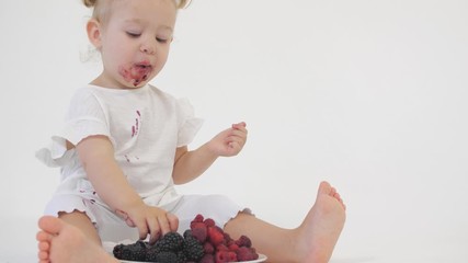Baby girl in white clothes eats juicy berries on light background
