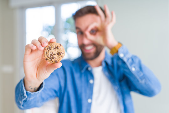 Handsome man eating chocolate chips cookies with happy face smiling doing ok sign with hand on eye looking through fingers