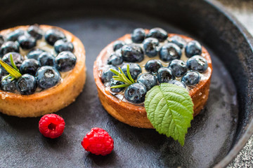 Delicious blueberry tartalets with vanilla cream and fresh raspberry berries on a black cast iron plate. 