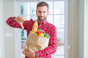 Handsome man holding groceries bag with angry face, negative sign showing dislike with thumbs down, rejection concept