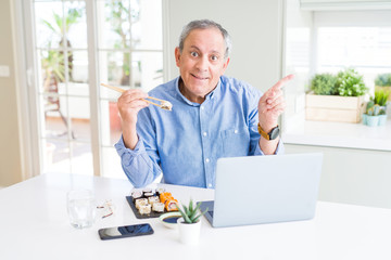 Handsome business senior man eating delivery sushi while working using laptop very happy pointing with hand and finger to the side