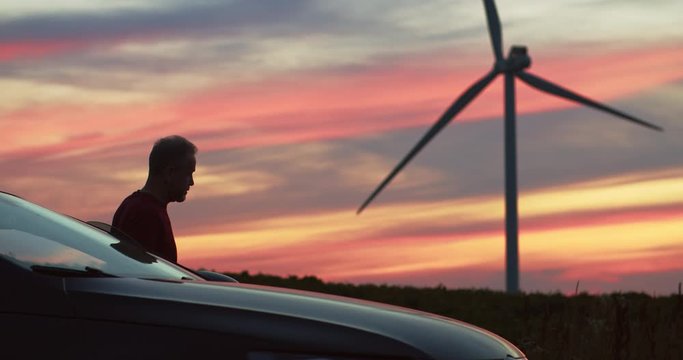 Handsome man arriving at windmill station admiring amazing sunset pink lights. Modern wind turbines clean power generators rotating in background.