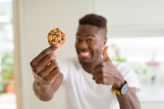 Young african american man eating chocolate chips cookies happy with big smile doing ok sign, thumb up with fingers, excellent sign