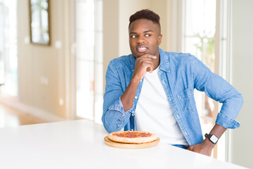 African american man eating pepperoni pizza at home serious face thinking about question, very confused idea