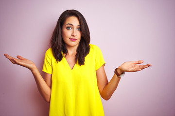 Young beautiful woman wearing yellow t-shirt standing over pink isolated background clueless and confused expression with arms and hands raised. Doubt concept.