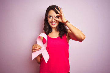 Young beautiful woman holding cancer ribbon over pink isolated background with happy face smiling doing ok sign with hand on eye looking through fingers