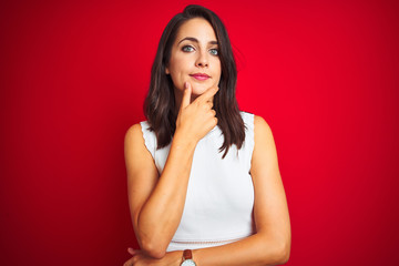 Young beautiful woman wearing white dress standing over red isolated background looking confident at the camera with smile with crossed arms and hand raised on chin. Thinking positive.