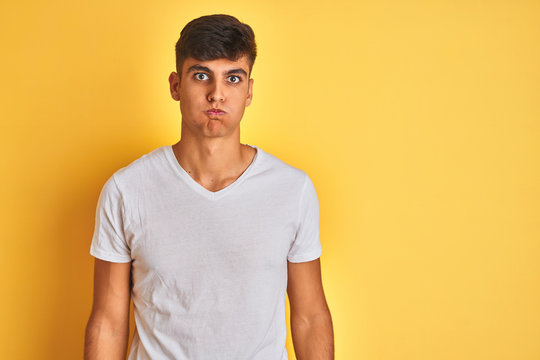 Young indian man wearing white t-shirt standing over isolated yellow background puffing cheeks with funny face. Mouth inflated with air, crazy expression.