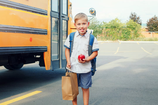 Funny cute happy Caucasian boy student kid with backpack, red apple and brown paper bag near yellow bus on 1 September day. Education and back to school concept. Child making silly faces.