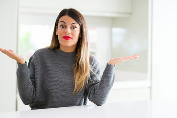 Young beautiful woman wearing winter sweater at home clueless and confused expression with arms and...