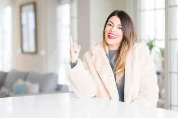Obraz premium Young beautiful woman wearing winter fur coat at home with a big smile on face, pointing with hand and finger to the side looking at the camera.