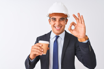 Young handsome architect man wearing helmet drinking coffee over isolated white background doing ok sign with fingers, excellent symbol