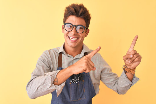 Young handsome hairdresser man wearing apron and glasses over isolated yellow background smiling and looking at the camera pointing with two hands and fingers to the side.