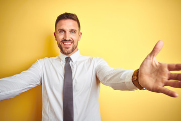 Young handsome business man wearing elegant white shirt over yellow isolated background looking at the camera smiling with open arms for hug. Cheerful expression embracing happiness.