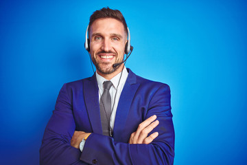 Young handsome operator man wearing call center headset over blue isolated background happy face smiling with crossed arms looking at the camera. Positive person.