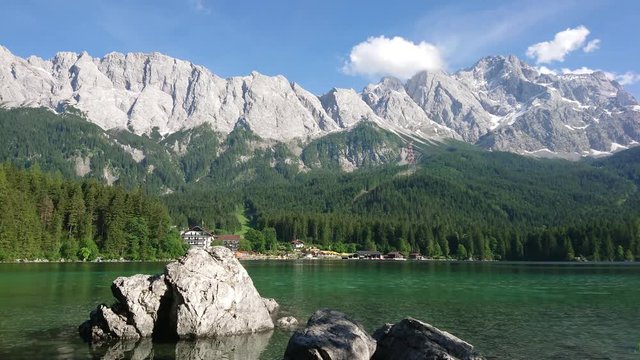 Spectacular view of the huge rocky and snowy mountain chain with green trees at slope, cloudy blue sky at the background and rocks in clear lake water on foreground on a sunny bright afternoon.