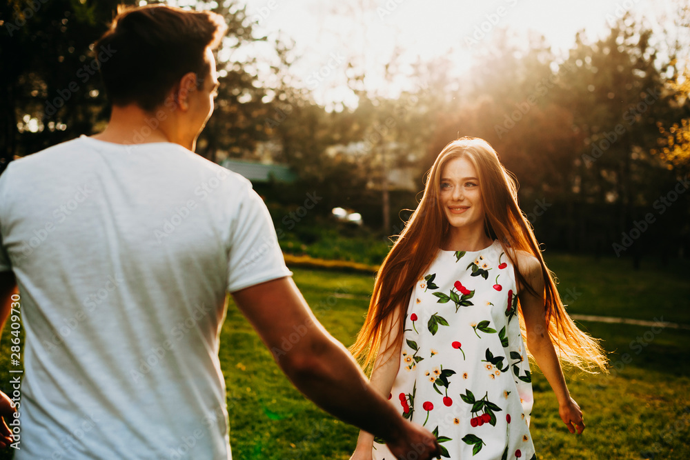 Sticker Charming woman with long red hair and freckles smiling and looking at her boyfriend while dancing in the wind outside against sunset and trees.