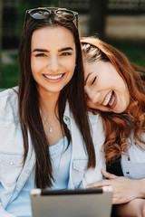 Two lovely caucasian girlfriend having fun while sitting on a bench looking at camera laughing with holding a tablet.