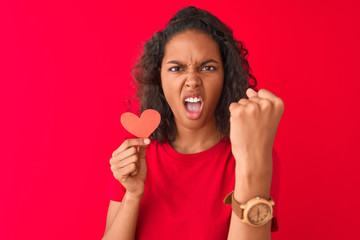 Young brazilian woman holding paper heart standing over isolated red background annoyed and frustrated shouting with anger, crazy and yelling with raised hand, anger concept