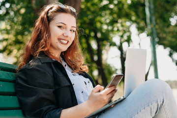 Portrait of a charming plus size woman sitting on a bench with a laptop on her legs and a smartphone in one hand looking at camera smiling.
