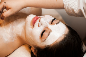 Close up portrait of a charming brunette leanin on a bed with closed eyes having a skin care routine with white mask in a wellness center.