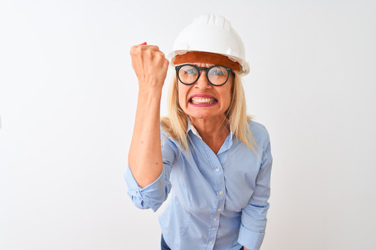 Middle age architect woman wearing glasses and helmet over isolated white background angry and mad raising fist frustrated and furious while shouting with anger. Rage and aggressive concept.