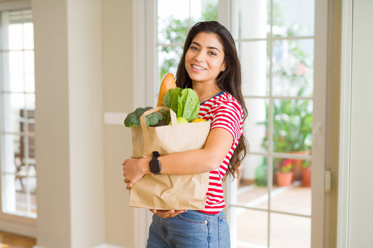 Beautiful young woman smiling holding a paper bag full of fresh groceries at home