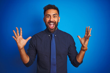Young indian businessman wearing elegant shirt and tie standing over isolated blue background celebrating crazy and amazed for success with arms raised and open eyes screaming excited. Winner concept