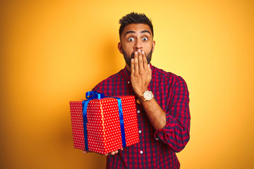 Arab indian hispanic man holding birthday gift standing over isolated yellow background cover mouth with hand shocked with shame for mistake, expression of fear, scared in silence, secret concept