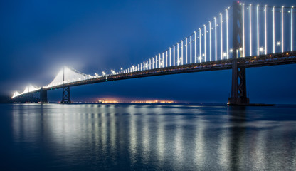 Fototapeta na wymiar San Francisco Bay Bridge at night with deep blue sky and hazy lights in the fog