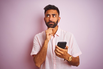 Young indian man using smartphone standing over isolated pink background serious face thinking...