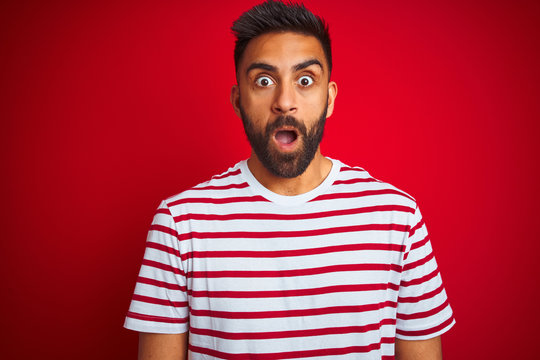 Young indian man wearing striped t-shirt standing over isolated red background afraid and shocked with surprise expression, fear and excited face.