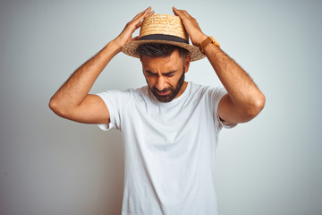 Young indian man on holiday wearing summer hat standing over isolated white background suffering from headache desperate and stressed because pain and migraine. Hands on head.