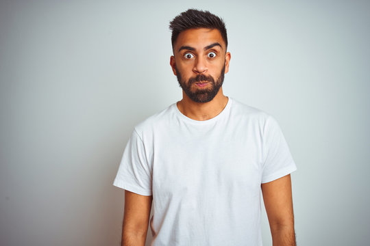 Young indian man wearing t-shirt standing over isolated white background puffing cheeks with funny face. Mouth inflated with air, crazy expression.