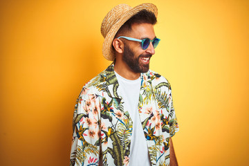 Indian man on vacation wearing floral shirt hat sunglasses over isolated yellow background looking away to side with smile on face, natural expression. Laughing confident.