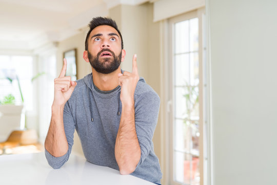 Handsome hispanic man wearing casual sweatshirt at home amazed and surprised looking up and pointing with fingers and raised arms.