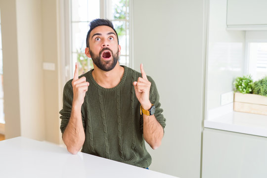 Handsome hispanic man wearing casual sweater at home amazed and surprised looking up and pointing with fingers and raised arms.