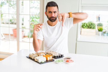 Handsome hispanic man eating asian sushi using chopsticks with angry face, negative sign showing dislike with thumbs down, rejection concept